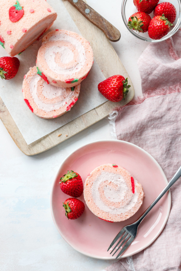 OVerhead shot of a strawberry cake roll, and a slice on a pink plate with fresh strawberries on the side.