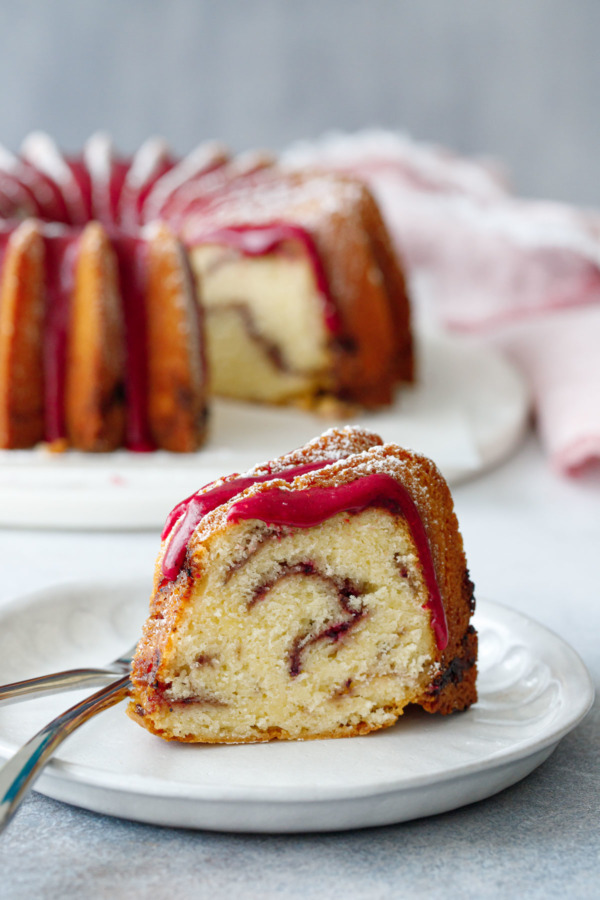 A slice of strawberry hibiscus swirl pound cake sitting on a plate, with the full bundt cake on a marble cake plate in the background.