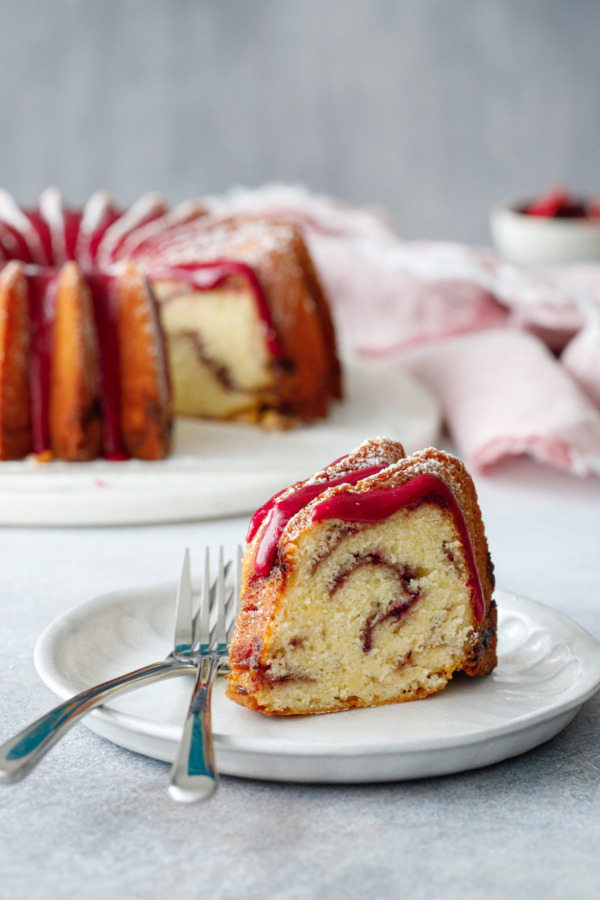 A slice of strawberry hibiscus swirl pound cake sitting on a plate, with the full bundt cake on a marble cake plate in the background.