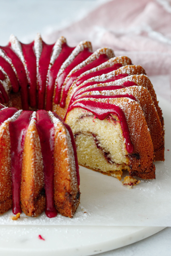 A brilliant bundt cake with a slice taken out of it, dusted in powdered sugar with a bright pink hibiscus glaze.