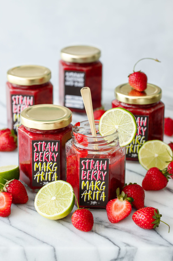 Five jars of Strawberry Margarita Jam on a marble surface, surrounded by fresh strawberries and limes.