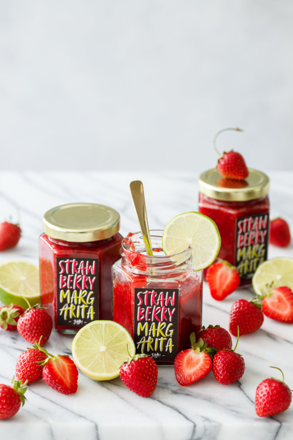 Three jars of homemade Strawberry Margarita Jam, one jar open with a gold spoon and a slice of lime on the edge, surrounded by fresh strawberries and limes.