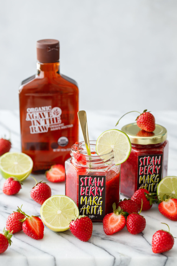 Two jars of Strawberry Margarita Jam with a bottle of organic agave syrup and fresh strawberries and limes.