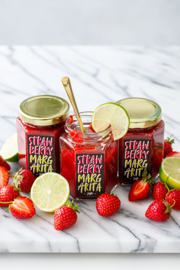 Three jars of homemade Strawberry Margarita Jam with custom-designed printable labels, one jar open with a gold spoon.