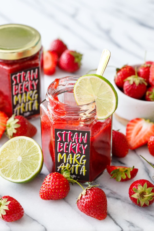 Homemade strawberry margarita jam in hexagon glass jars with custom designed labels, surrounded by fresh strawberries and limes.