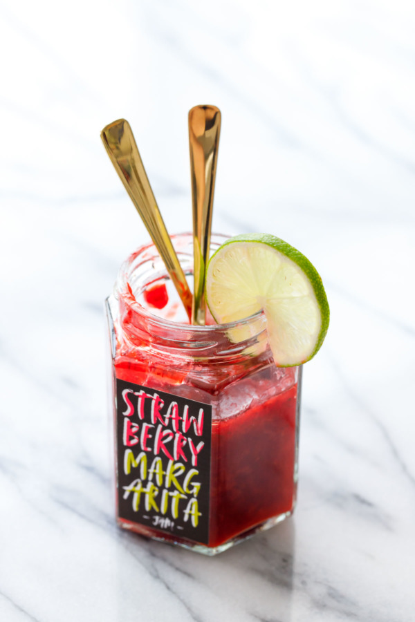 Hexagon glass jar filled with bright red Strawberry Margarita Jam, with two gold spoons and a slice of lime on the rim of the jar.