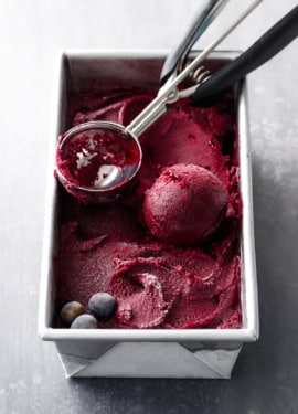 A metal loaf pan filled with Blueberry Ginger Sorbet, with an ice cream scoop and a perfect ball of sorbet.