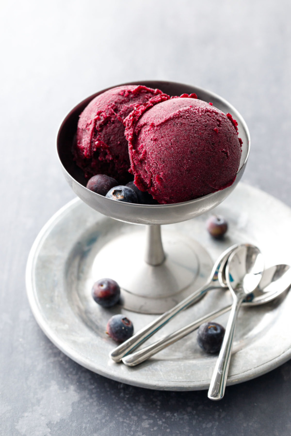 A metal ice cream pedestal dish with two scoops of Blueberry Ginger Sorbet, on a pewter plate with silver spoons.