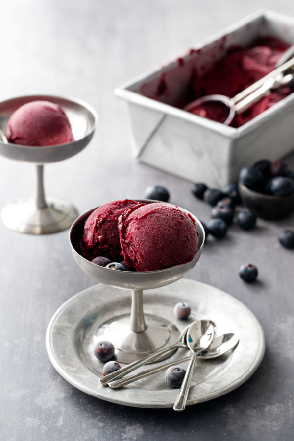 Two scoops of Blueberry Ginger Sorbet in a stainless steel ice cream dish, with a another scoop and a pan full of sorbet in the background.