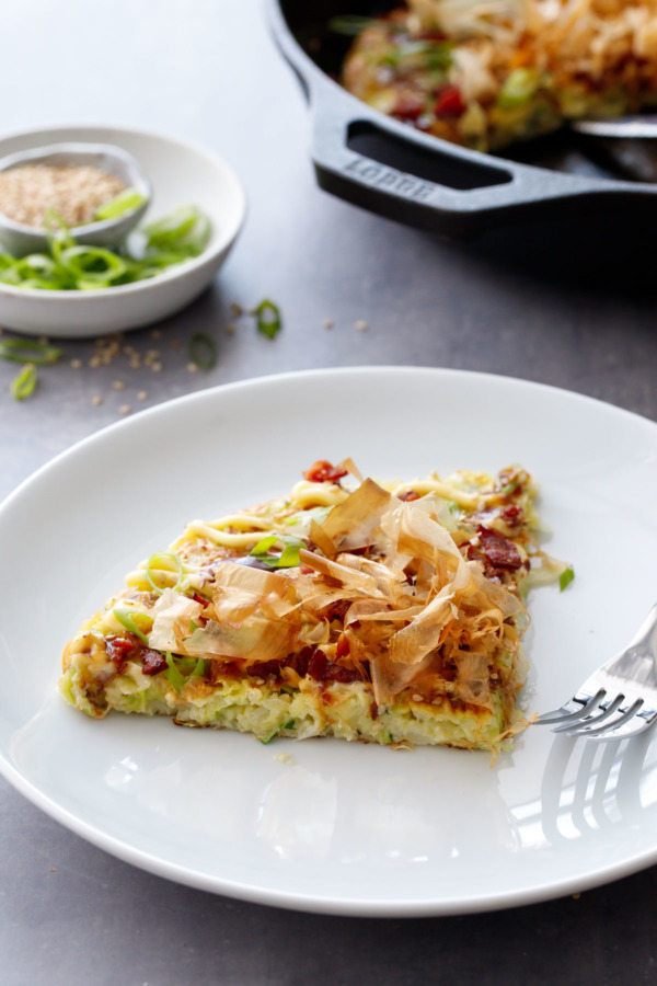 A slice of Cabbage & Zucchini Okonomiyaki on a plate, with a cast iron skillet in the background