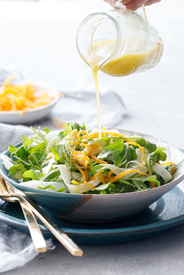 Drizzling an orange vinaigrette dressing onto a bowl of arugula fennel salad, dish of candied orange peel in the background.