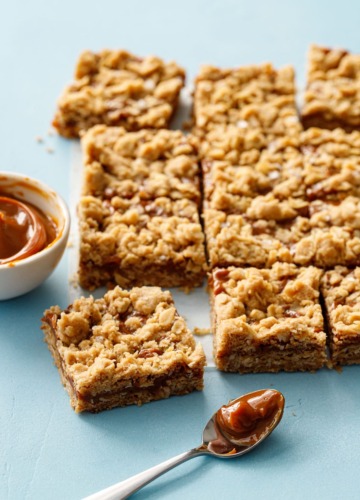 Dulce de Leche Oatmeal Crumb Bars on a blue background with a bowl and spoon of dulce de leche