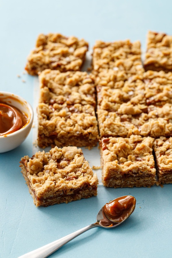 Dulce de Leche Oatmeal Crumb Bars on a blue background with a bowl and spoon of dulce de leche