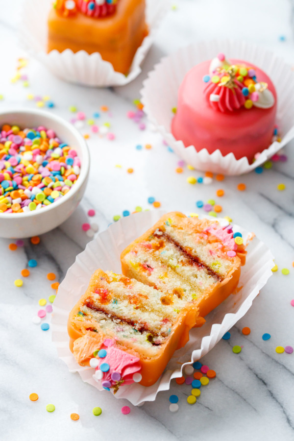 Orange-glazed "Funfetti Fours" cut in half to show the layers of funfetti cake, apricot and raspberry jams.