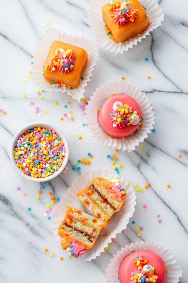 Overhead view of pink and orange "funfetti fours", one cake cut in half to show the apricot and raspberry jam layers, with a bowl of rainbow sprinkles.