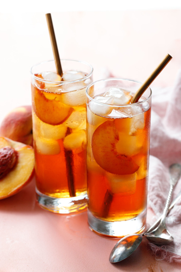 Two glasses filled with Cold Brew Peach Iced Tea, with rose gold metal drinking straws and fresh peaches on the side.