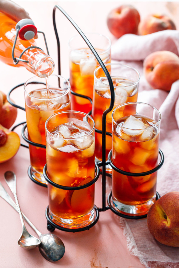 A vintage drink caddy with 5 glasses of Cold Brew Peach Iced Tea, and one bottle of peach sugar syrup being poured into one of the glasses.