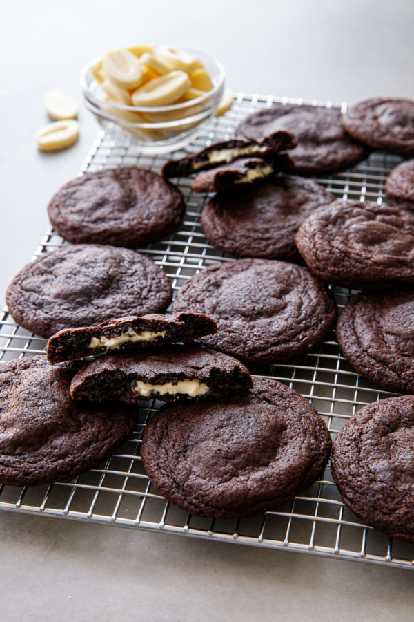 Wire rack with chocolate cookies arranged randomly, two cookies broken in half to show the white chocolate filling, with a bowl of white chocolate pieces.