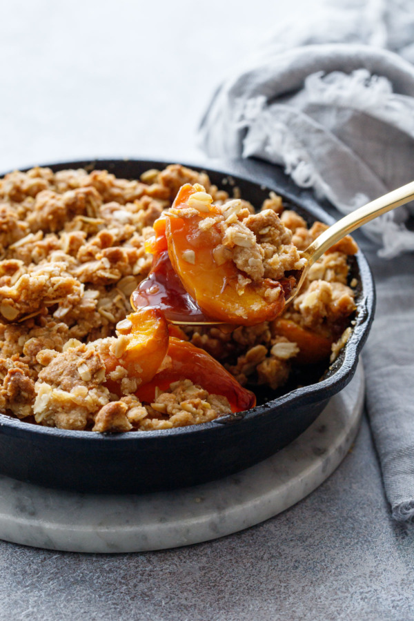 A serving spoon scooping a heaping serving of Skillet Bourbon Peach Crisp out of a cast iron skillet