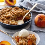 Skillet Bourbon Peach Crisp with fresh peaches on the side and a shot glass of bourbon.