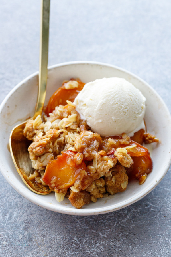 Small bowl with a scoop of Bourbon Peach Crisp and a scoop of vanilla ice cream with a gold spoon.