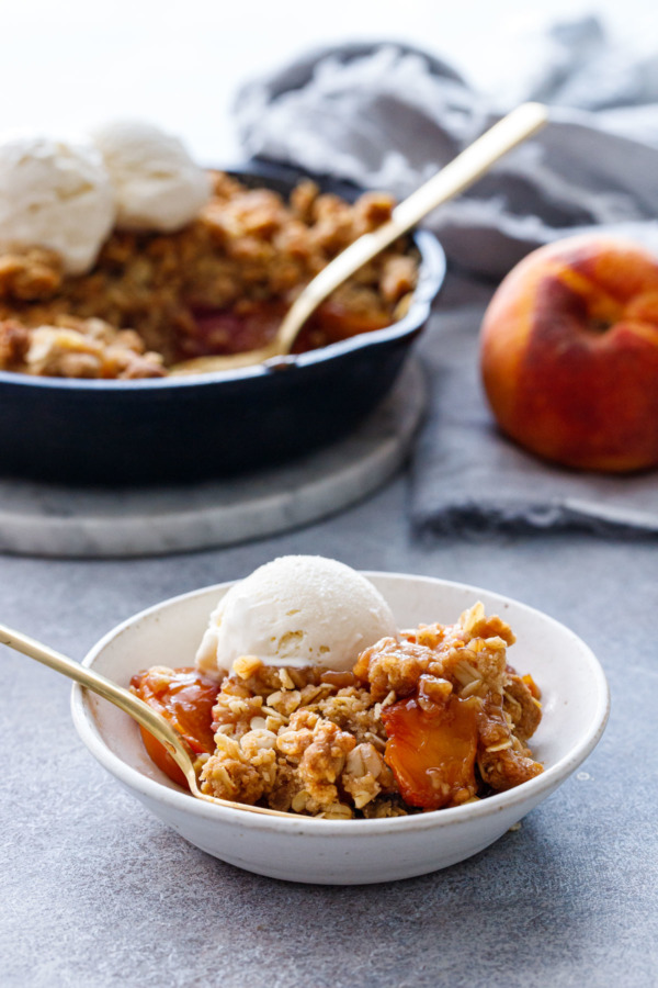 A smal bowl with a scoop of Bourbon Peach Crisp and vanilla ice cream, with fresh peaches and a cast iron skillet in the background.