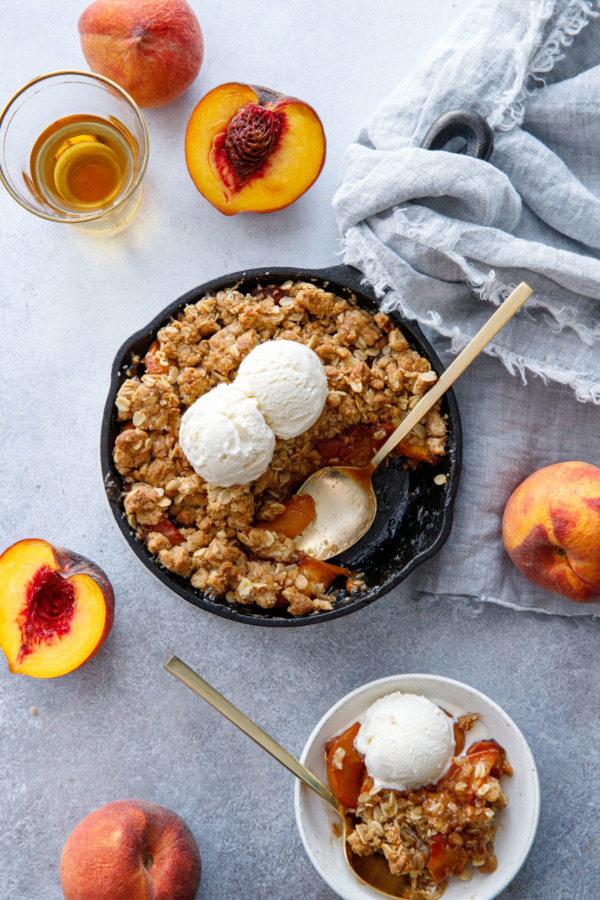 Overhead shot of Skillet Bourbon Peach Crisp on a gray background, with fresh peaches and a shot glass of bourbon.