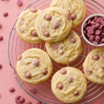 Overhead shot of Cacao Butter and Ruby Chocolate Chip Cookies on a wire rack with a dish filled with ruby cacao wafers, pink background.