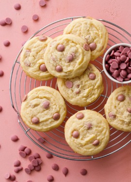 Overhead shot of Cacao Butter and Ruby Chocolate Chip Cookies on a wire rack with a dish filled with ruby cacao wafers, pink background.