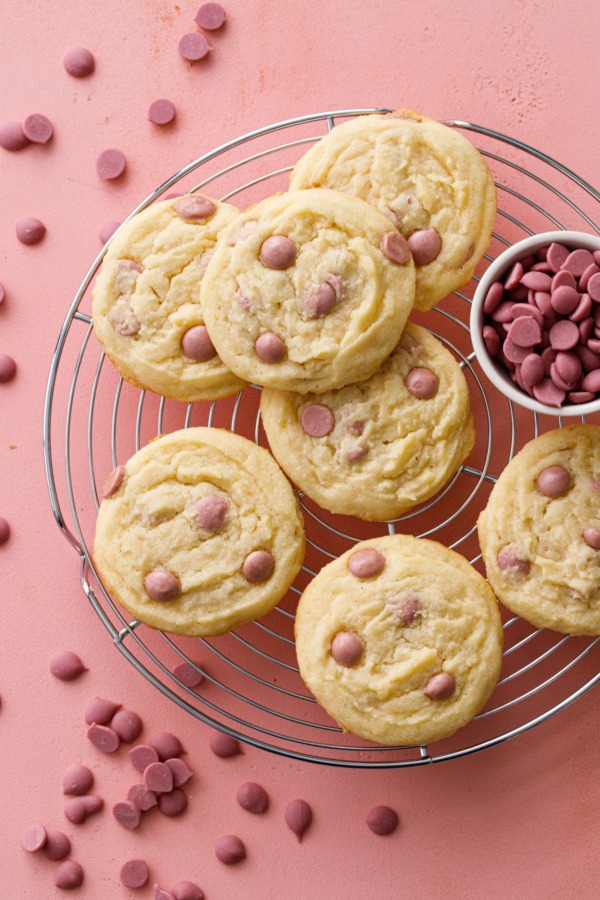 Overhead shot of Cacao Butter and Ruby Chocolate Chip Cookies on a wire rack with a dish filled with ruby cacao wafers, pink background.