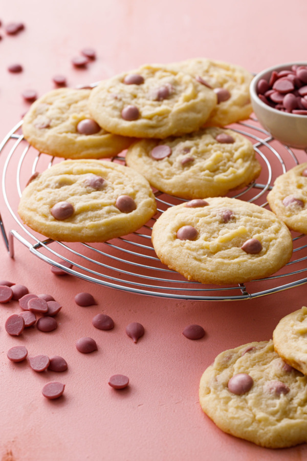 Cacao Butter and Ruby Chocolate Chip Cookies on a wire rack with ruby cacao wafers scattered around on a pink background.