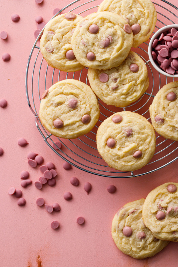 Overhead shot of Cacao Butter and Ruby Chocolate Chip Cookies on a wire rack with ruby cacao wafers scattered around on a pink background.