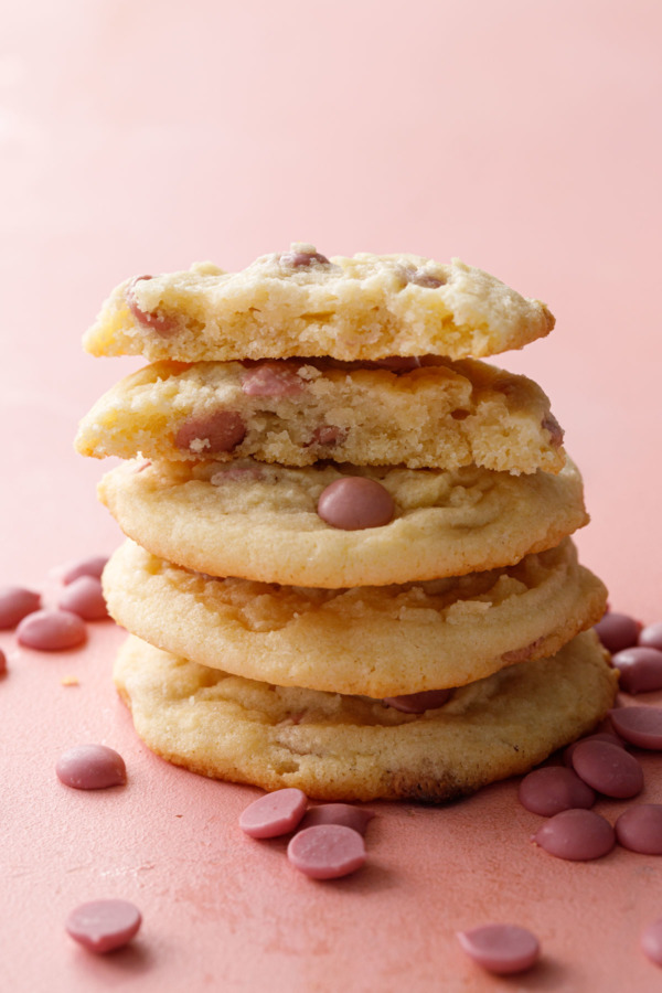 Stack of Cacao Butter and Ruby Chocolate Chip Cookie on a pink background, one cookie broken in half to show the texture inside.