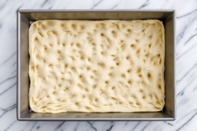 How to make homemade focaccia bread: press dough out to fill the baking pan, digging your fingers into the dough to give it its characteristic dimples.