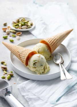 Two cones of pistachio gelato overturned on a white plate with two spoons and dish of green pistachios on the side.