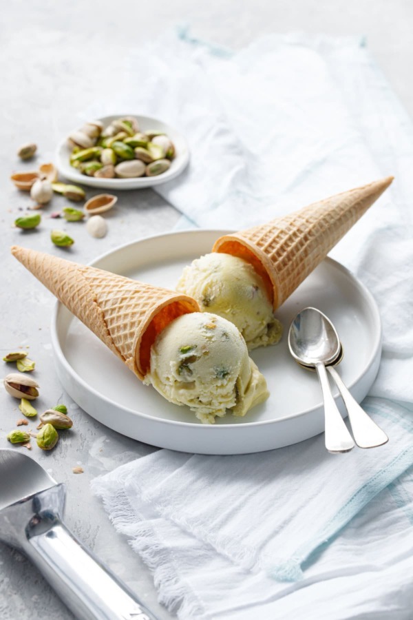 Two cones of pistachio gelato overturned on a white plate with two spoons and dish of green pistachios on the side.