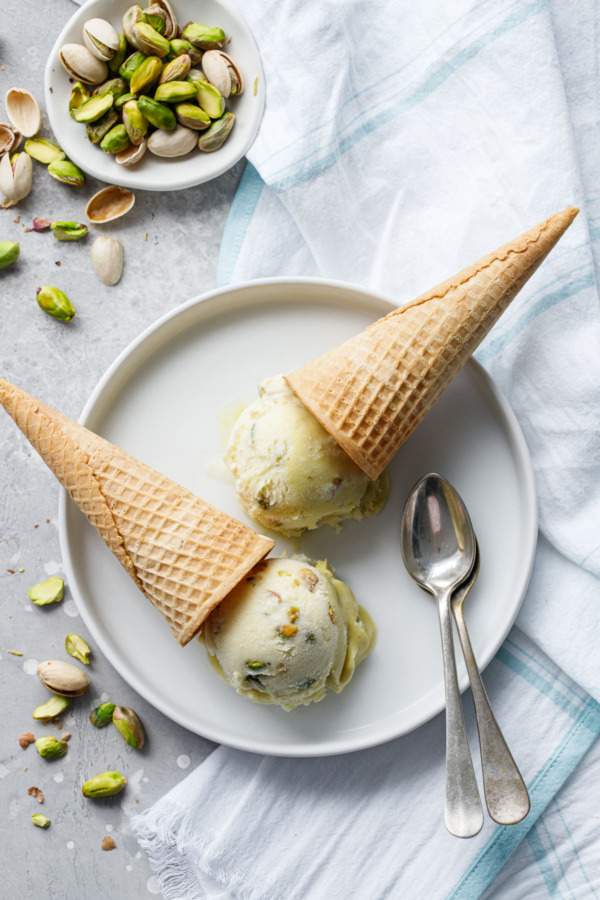 Overhead shot showing two cones of pistachio gelato overturned on a white plate with two spoons and dish of green pistachios on the side.
