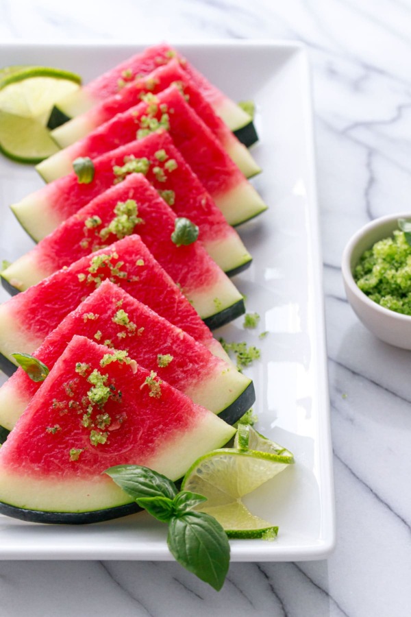 White plate with slices of watermelon, sprinkled with basil lime sugar. Garnished with fresh lime slices and a small bowl of sugar on the side.