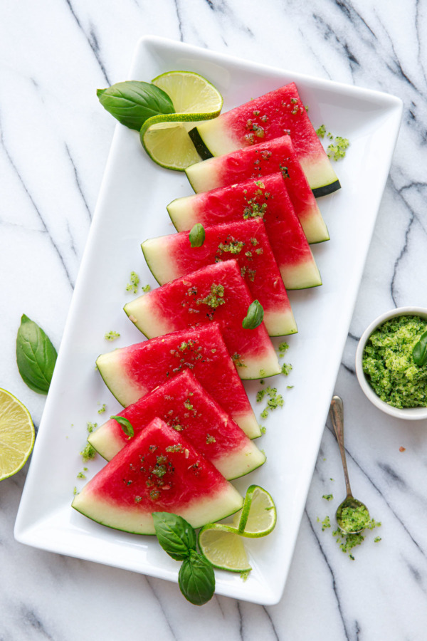 Overhead shot of watermelon slices overlapping on a rectangular white plate, sprinkled with basil lime sugar and a small bowl and spoon of sugar on the side.