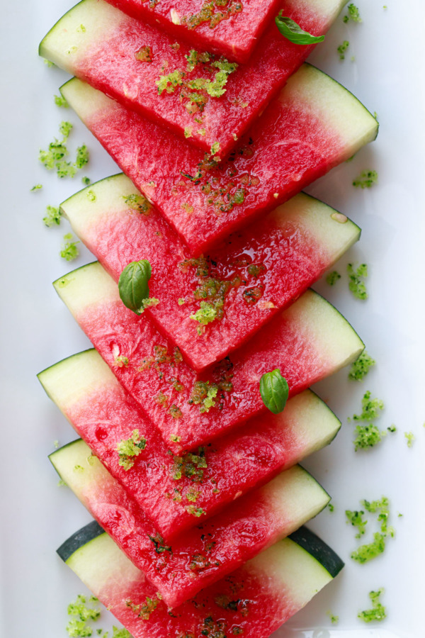 Triangular slices of seedless red watermelon overlapping on a white plate, sprinkled with basil lime sugar
