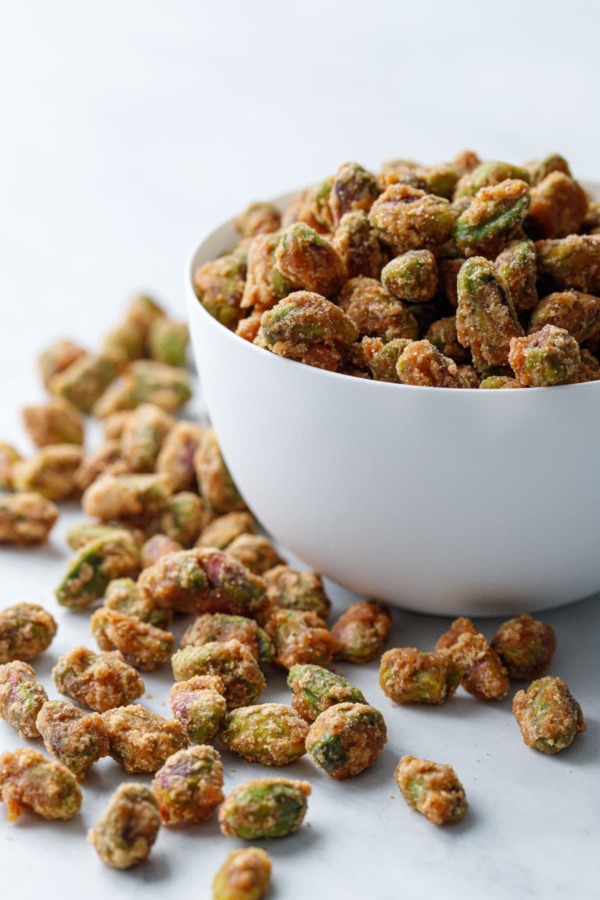 A white porcelain bowl overflowing with spiced candied pistachios on a marble surface