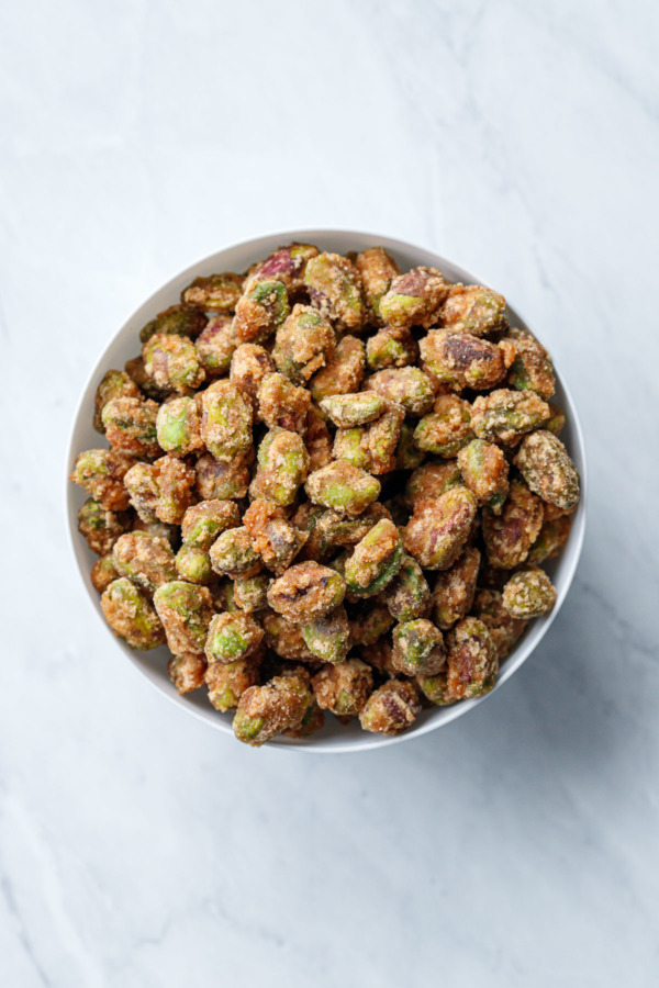 Overhead view of a white porcelain bowl filled with sweet & spicy crystalized candied pistachios, on a marble background.