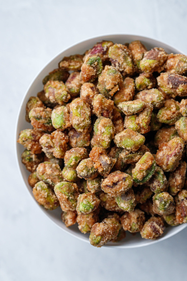 Close up overhead view of a white porcelain bowl filled with sweet & spicy crystalized candied pistachios, on a marble background.