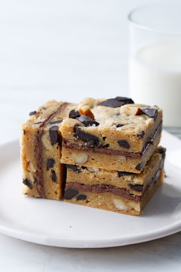 Three Nutella-Stuffed Brown Butter Blondies stacked on a white plate with a glass of milk in the background