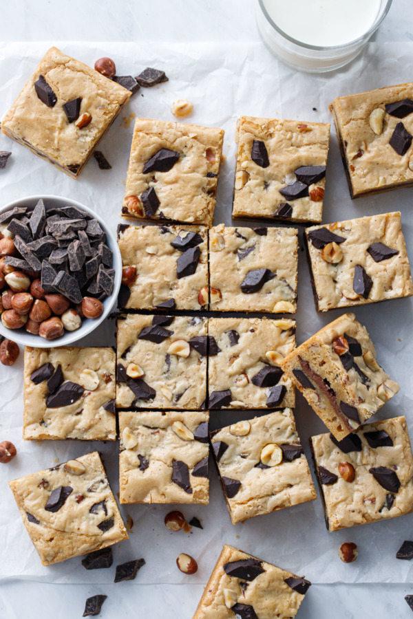 Overhead shot of Nutella-Stuffed Brown Butter Blondies cut into squares with a bowl of chocolate chunks and hazelnuts on the side.