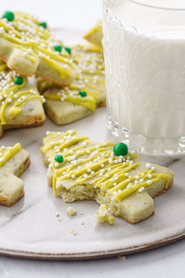 A pistachio Christmas tree cookie with a bite out of it, on a plate with a glass of milk in the background.