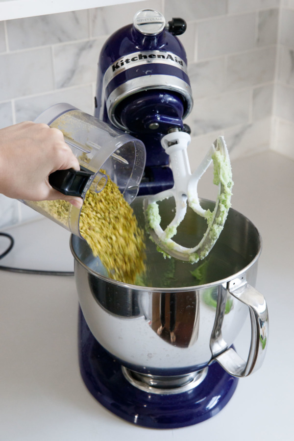 Pouring finely chopped pistachio cookies into the bowl of a stand mixer.