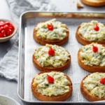 Cheesy Artichoke Crostini toasts arranged on a silver baking sheet with a bowl of artichoke topping and pepper drops on the side.