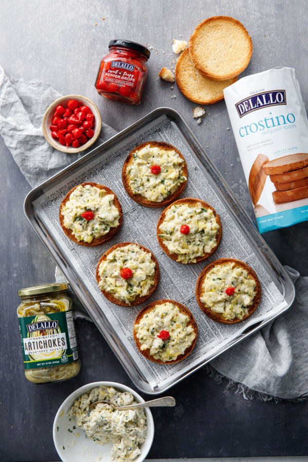 Overhead view of Cheesy Artichoke Crostini on a baking pan, with DeLallo's Crostino toasts, marinated artichoke hearts, and Pepper Drops used in the recipe.