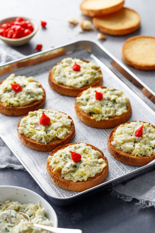 A silver baking pan with six artichoke crostino toasts topped with tiny red pepper drop peppers.